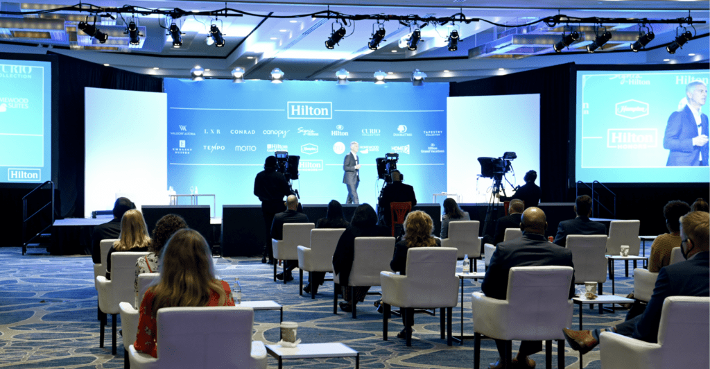 Audience watches a Hilton Hotels presentation from the floor of the hotel ballroom as a man presents onstage. 